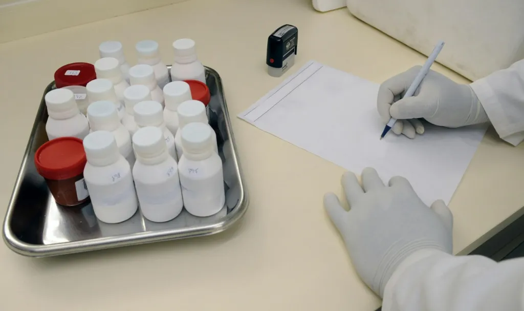 A gloved person writing on paper next to a tray of small white bottles.