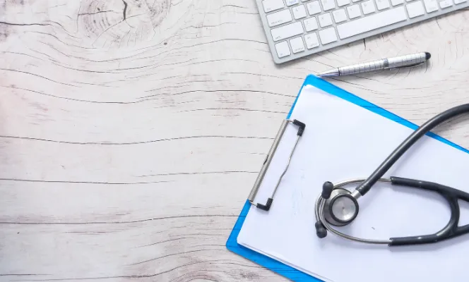 A clipboard with a blank paper and a black stethoscope resting on it, alongside a keyboard and pen on a wooden desk.