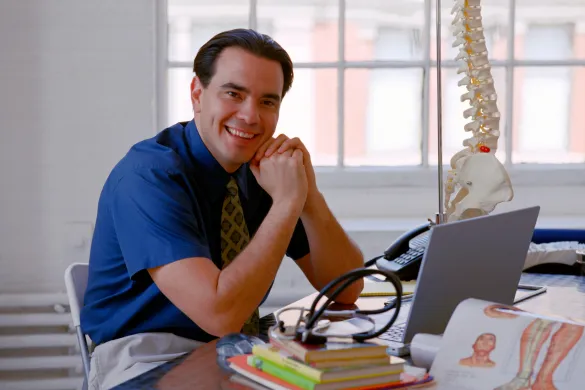 A smiling man sitting at a desk with a laptop, stethoscope, and a spine model in an office.