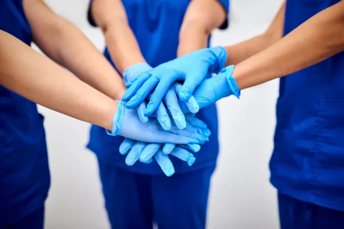 Healthcare workers in blue scrubs stacking their gloved hands together in unity.