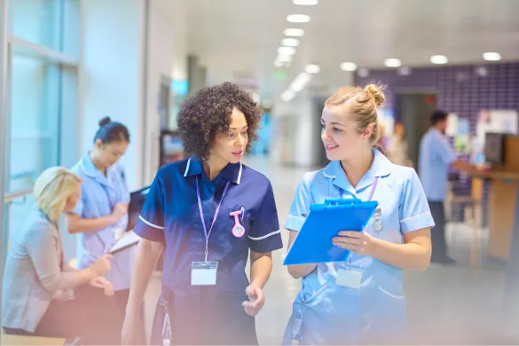 Two nurses walking and reviewing medical notes on a clipboard in a hospital hallway.