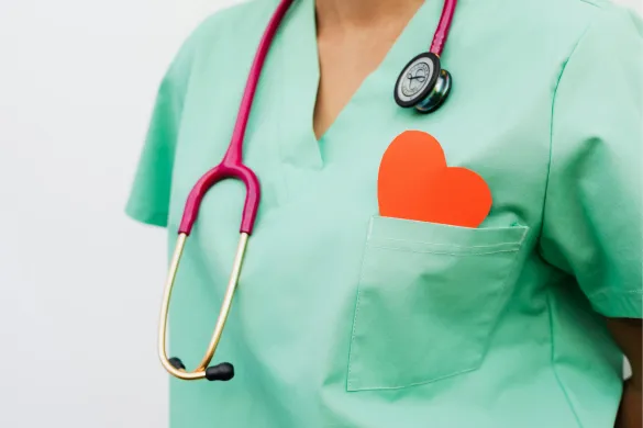 A healthcare worker wearing green scrubs and a pink stethoscope, with a red heart shape tucked into the chest pocket.