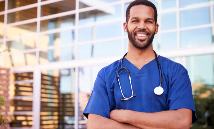 A smiling male healthcare worker in blue scrubs with a stethoscope standing outside a modern building.
