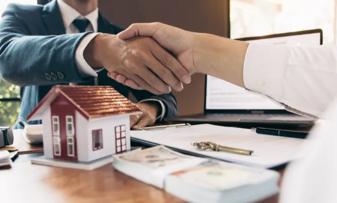 Two people shaking hands over a desk with a small model house, keys, and documents.
