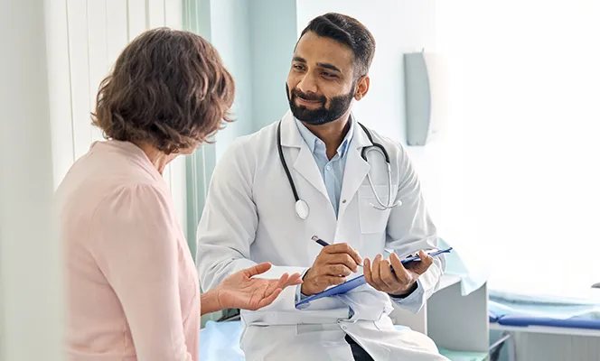 A smiling doctor talking with a patient while writing notes on a clipboard in an exam room.