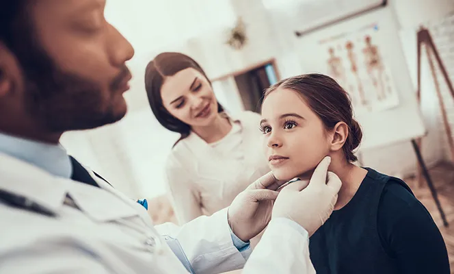 A pediatrician examining a young girl's throat while her mother watches in the background.
