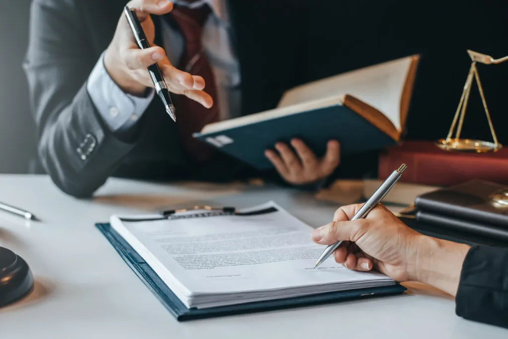 A lawyer gesturing while a client signs a legal document at a desk with law books and scales of justice.