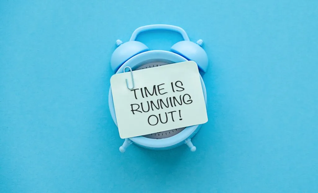 A light blue alarm clock on a matching blue background, featuring a sticky note attached with a paperclip that reads "TIME IS RUNNING OUT!".