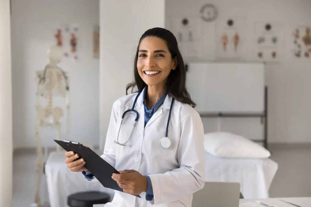 A smiling female doctor in a white lab coat holding a clipboard in a medical office.