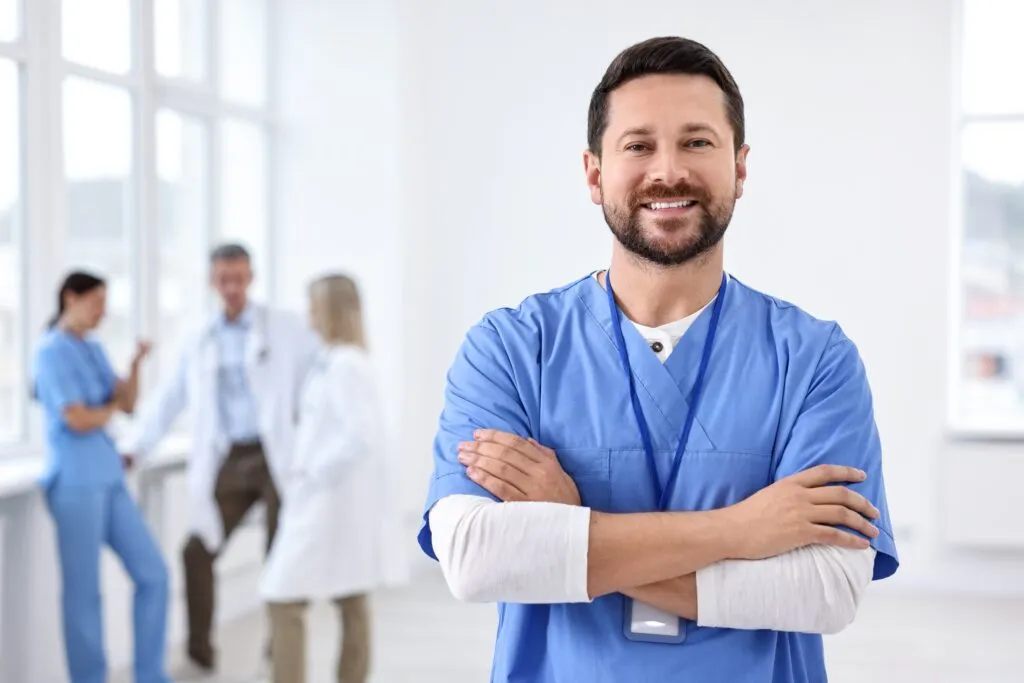 A smiling male nurse in blue scrubs standing with arms crossed, with colleagues talking in the background.