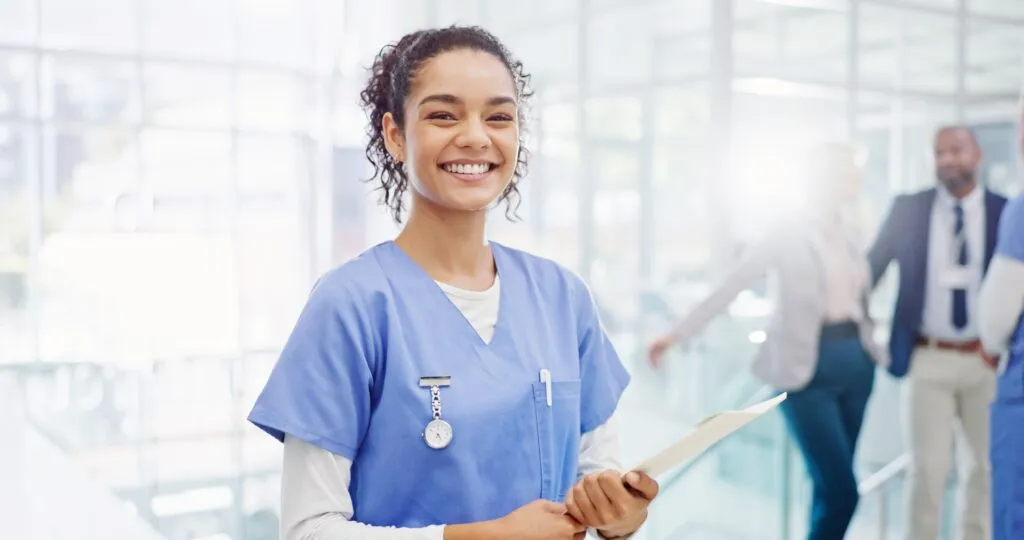 A smiling healthcare professional wearing blue scrubs and holding a clipboard.