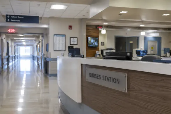 A modern hospital corridor featuring a "Nurses Station" desk in the foreground and a long hallway with patient rooms in the background.