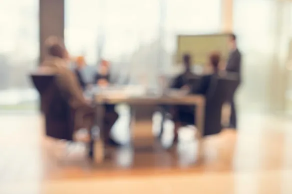 A blurred, soft-focus view of a group of professionals seated around a conference table in an office setting