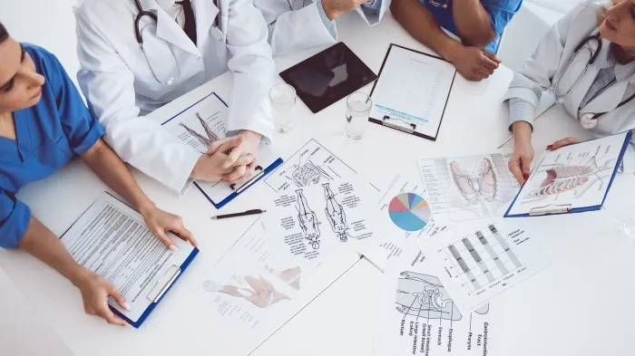 A top-down view of medical professionals gathered around a table reviewing charts, anatomical diagrams, and medical reports.